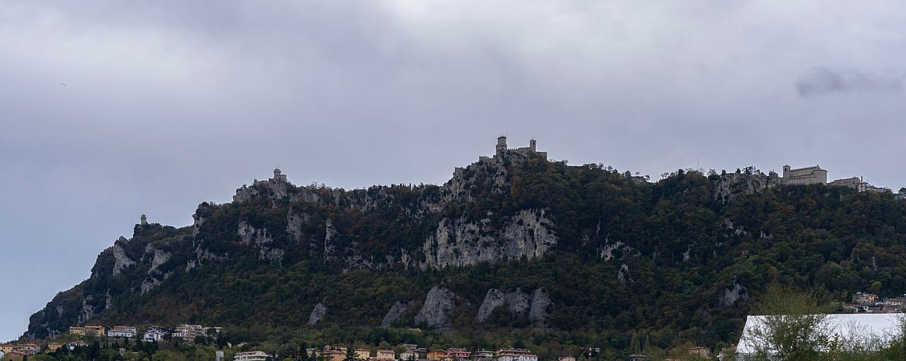 A panorama of Monte Titano, the limestone ridge that is the whole of San Marino, rising sharply from the Italian plain near the Adriatic. The capital clings to its upper slopes, crowned by the three towers that are the emblem of the nation.